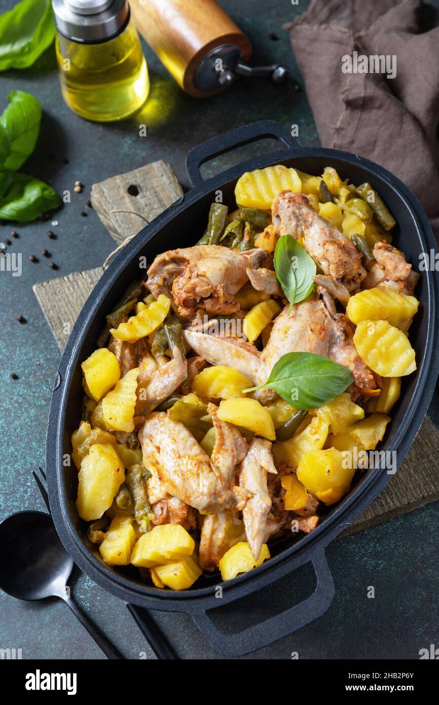 Gebackene Hähnchenflügel mit Kartoffeln, Bohnen, Kräutern, Knoblauch und Bazil auf einer Pfanne auf einer Tischplatte aus dunklem Stein. Hausgemachtes Essen. Griechische und mediterrane Küche. Stockfoto