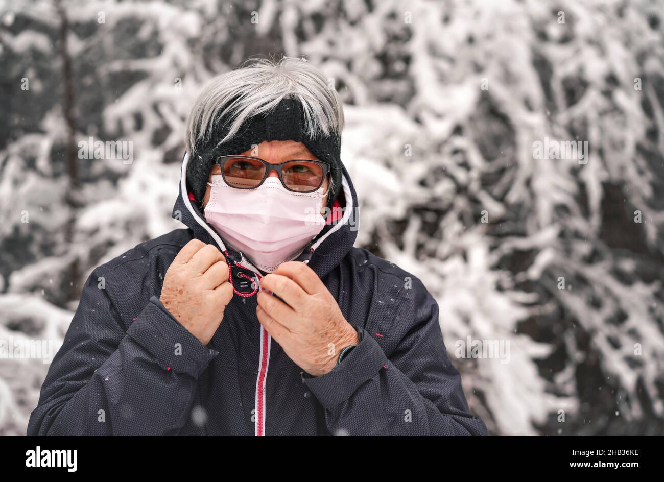 Ältere ältere Frau mit grauen Haaren, die eine rosa Einweg-Virus-Gesichtsmaske trägt, verschwommener Winter-Schnee bedeckte Bäume im Hintergrund Stockfoto