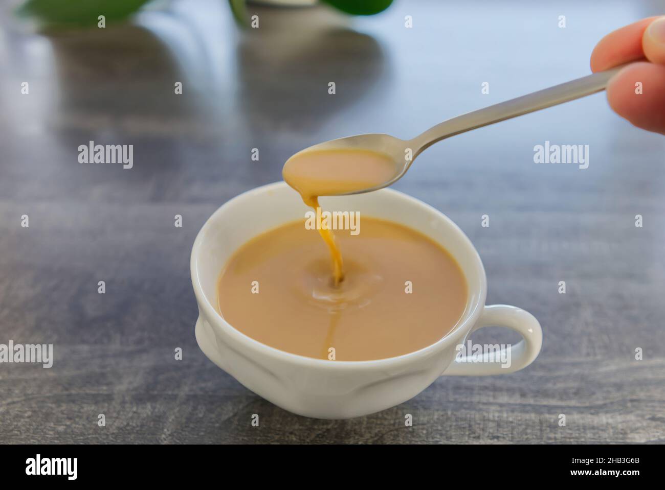 Kaffee mit Milch in einem weißen Becher. Die Hand rührt den Kaffee mit einem Löffel. Ein Tropfen Kaffee fällt in einen Becher. Stockfoto