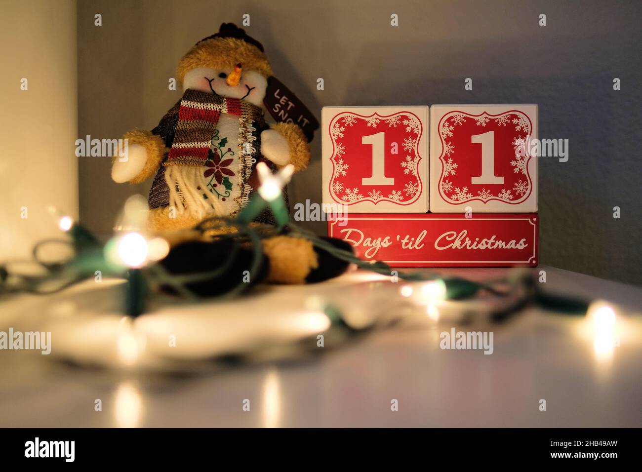 Elf Tage bis Weihnachten Countdown-Kalender aus Holzblöcken auf einem Mantel mit Lichtern und einem Plüsch Spielzeug Schneemann. Stockfoto