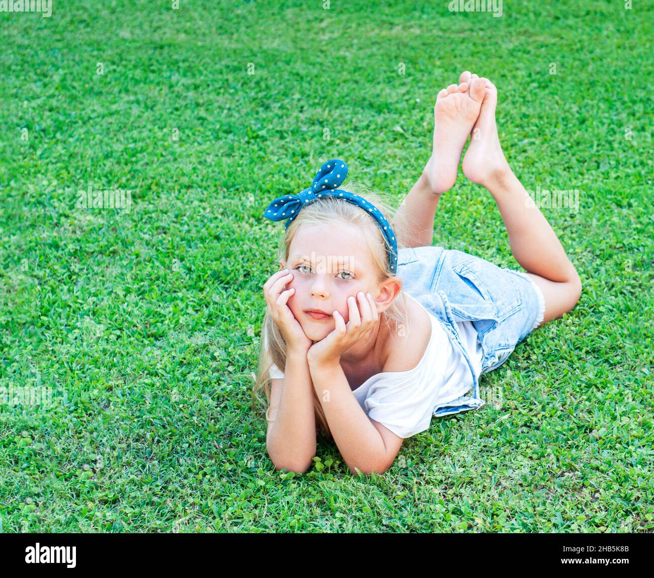 Сheerful kleines Mädchen in einem Jeansjumpsuit, auf einem grünen Gras liegend Stockfoto
