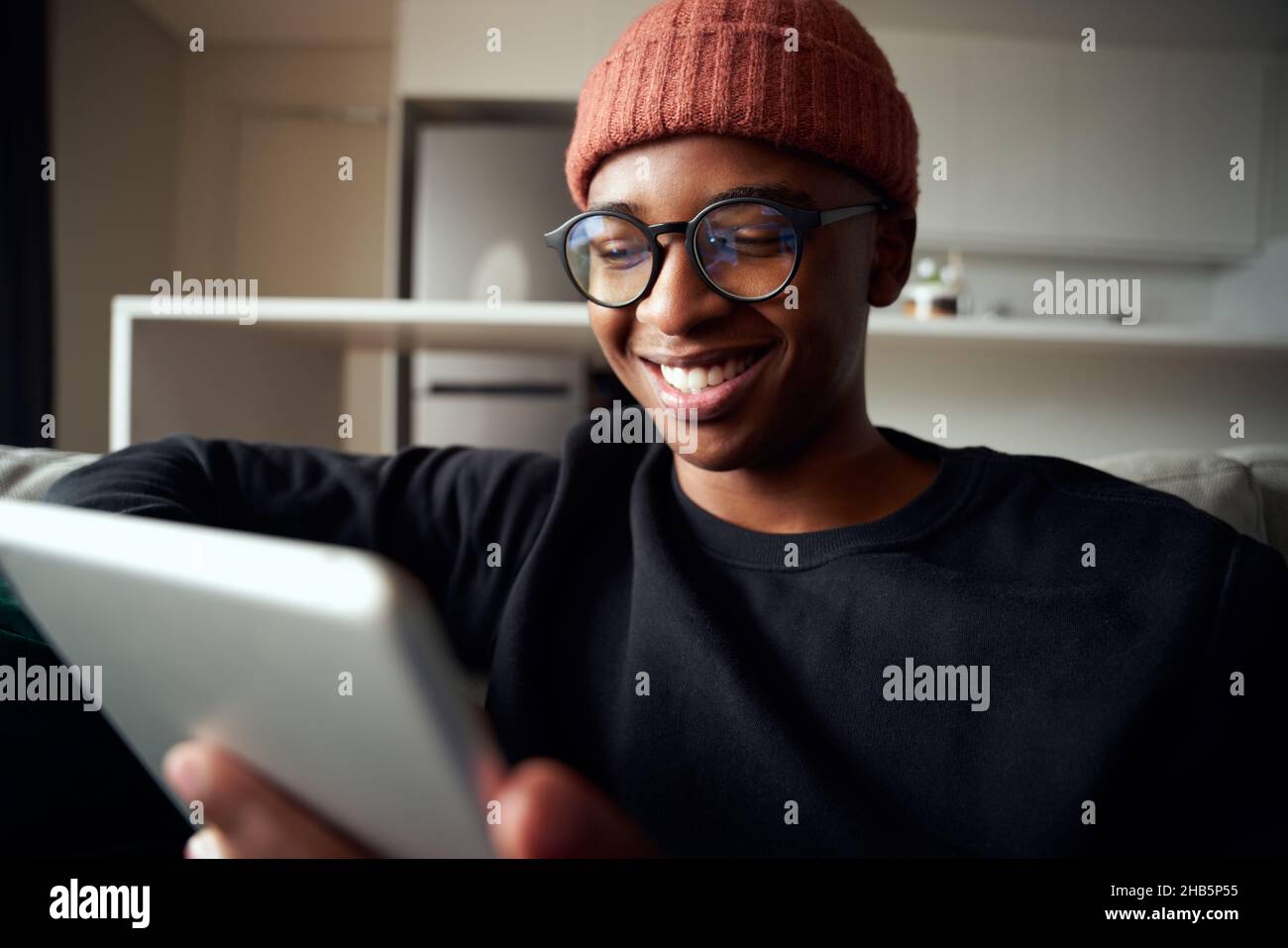 Nahaufnahme eines erwachsenen schwarzen Mannes, der lächelt, während er auf das Tablet schaut. Moderne Wohnung auf dem Sofa Stockfoto