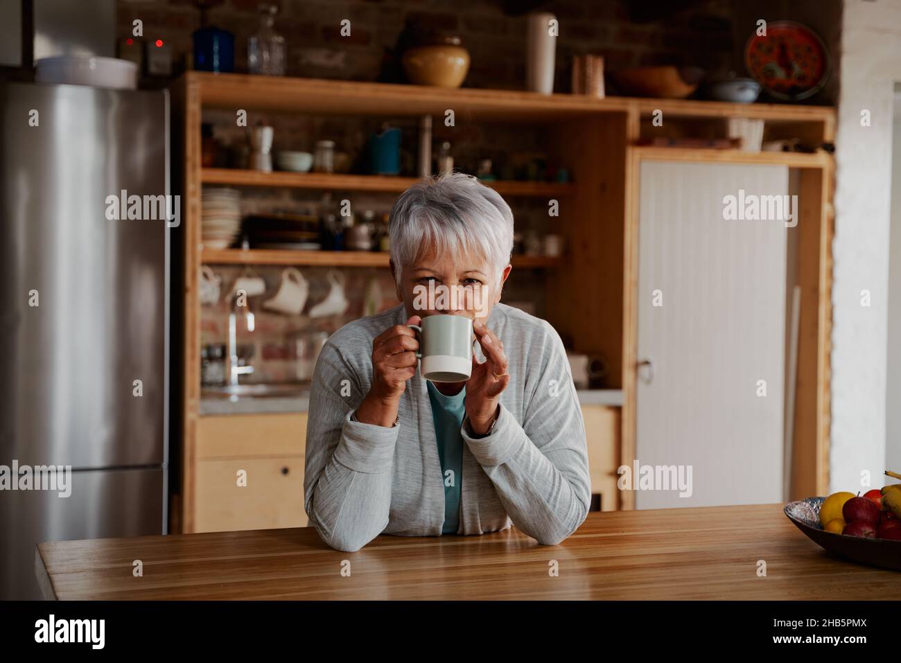 Porträt einer älteren Frau im Ruhestand, die sich auf der modernen Küchentheke stützt und die Kamera anlächelt und einen Schluck Kaffee am Morgen trinkt. Stockfoto