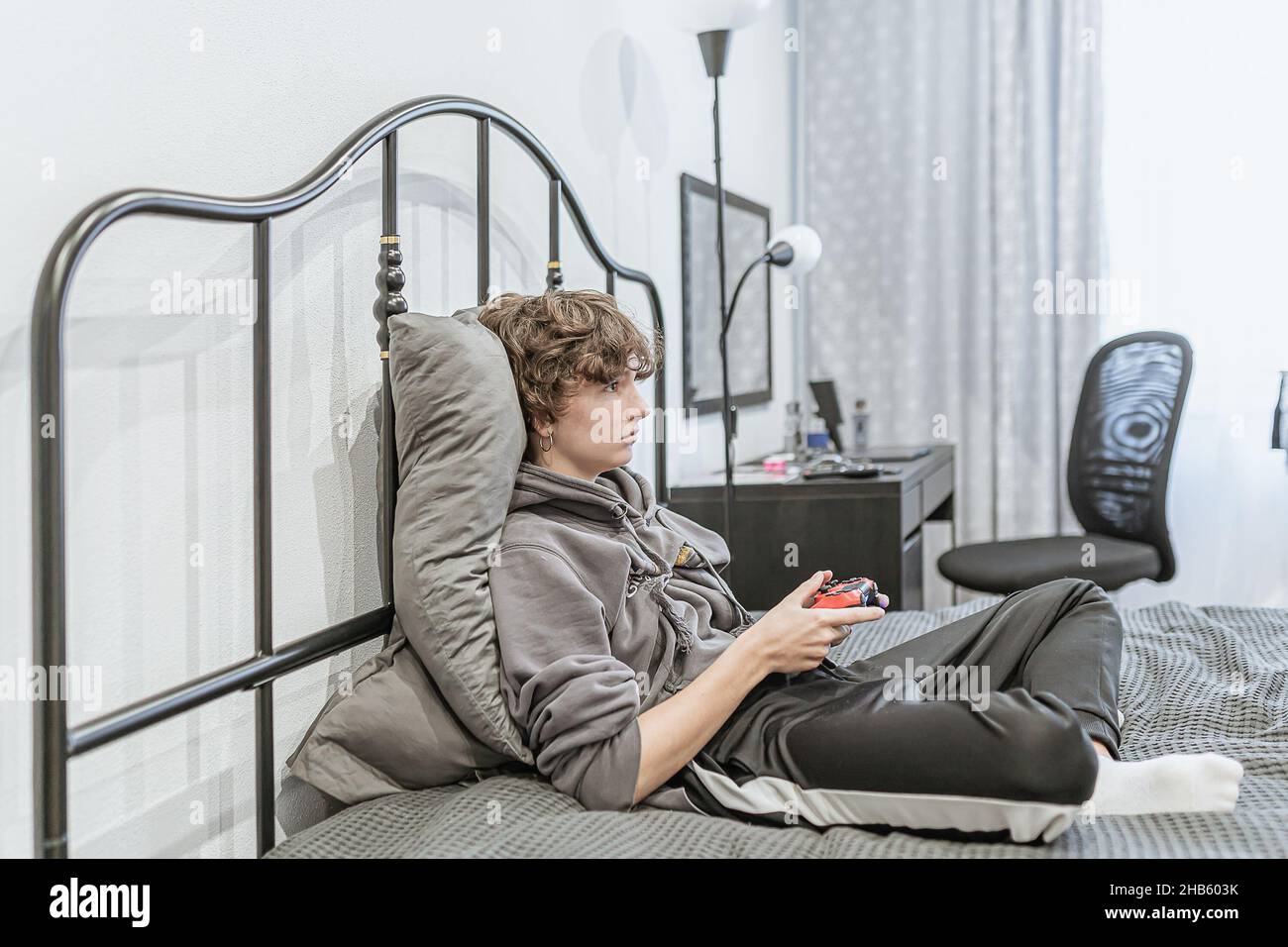 Teenager-Mädchen mit kurzen braunen lockigen Haaren, bekleidet mit ungepflegten Heimkleidung, sitzt auf einem ungefertigten Bett und spielt auf der Computerkonsole. Leidenschaft für Computer Stockfoto