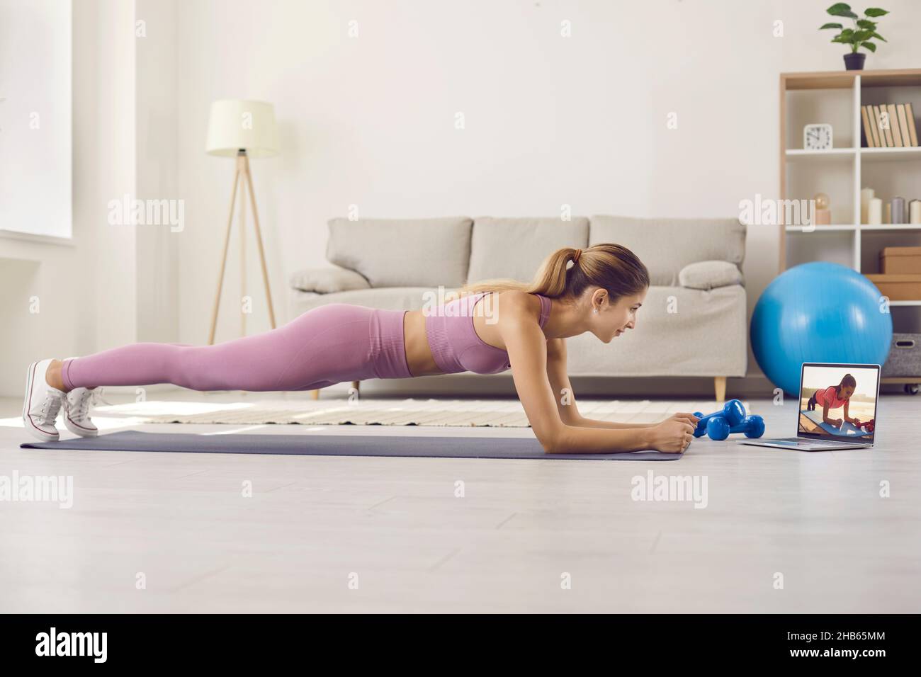Die junge, sportliche Frau arbeitet zu Hause und macht eine Planke vor ihrem Laptop. Stockfoto