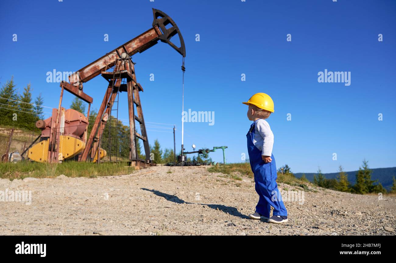 Kleiner Junge in blauem Overall und gelbem Kaské, der Offshore-Maschine in der Natur ansieht und über die Ausbildung von Technikern, das männliche Lernsystem von Bohrgerät und Bergbaupumpe nachdenkt Stockfoto