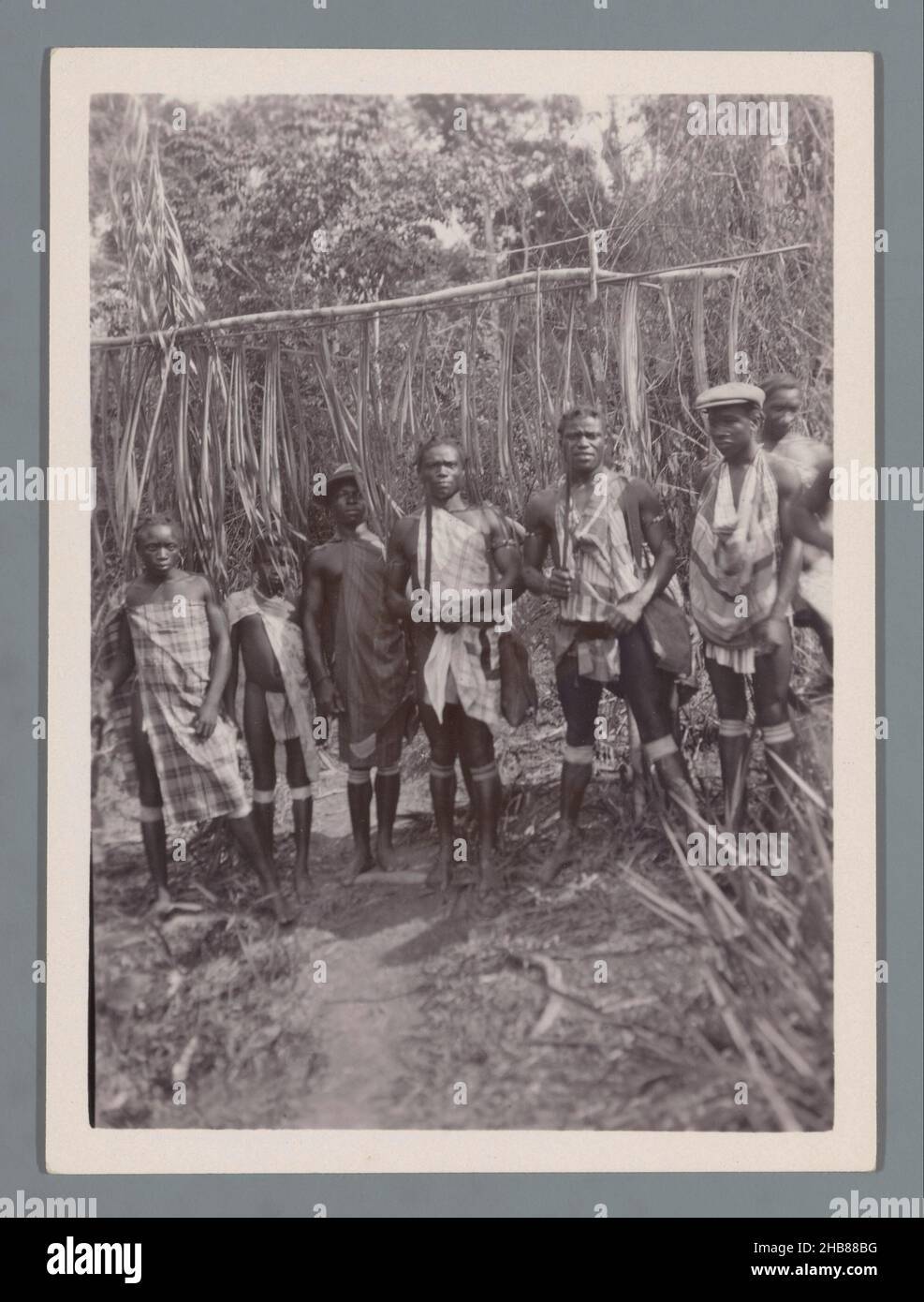 Sieben Maroon-Männer vor dem Eingang eines Maroon-Dorfes (vermutlich) in Suriname, anonym, c. 1910 - c. 1940, fotografischer Träger, Silbergelatine-Druck, Höhe 120 mm × Breite 88 mm Stockfoto