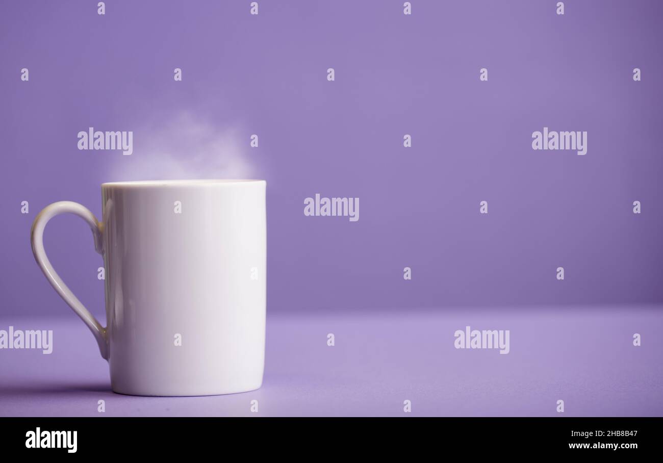 Weiße Tasse Kaffee oder Tee aus Keramik mit heißem Dampf, mit violettem Hintergrund (sehr peri). Stockfoto