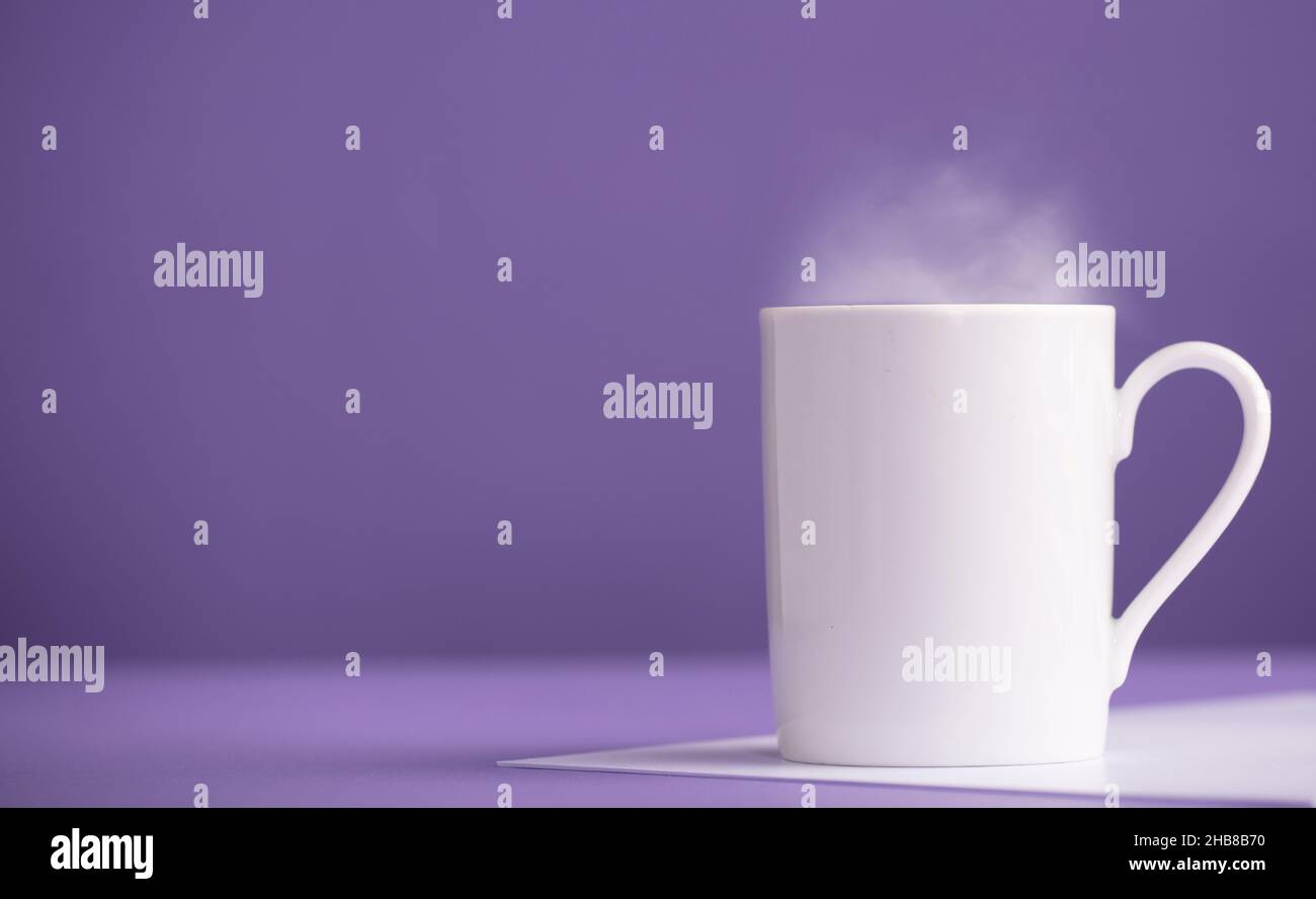 Weiße Tasse Kaffee oder Tee aus Keramik mit heißem Dampf, mit violettem Hintergrund (sehr peri). Stockfoto
