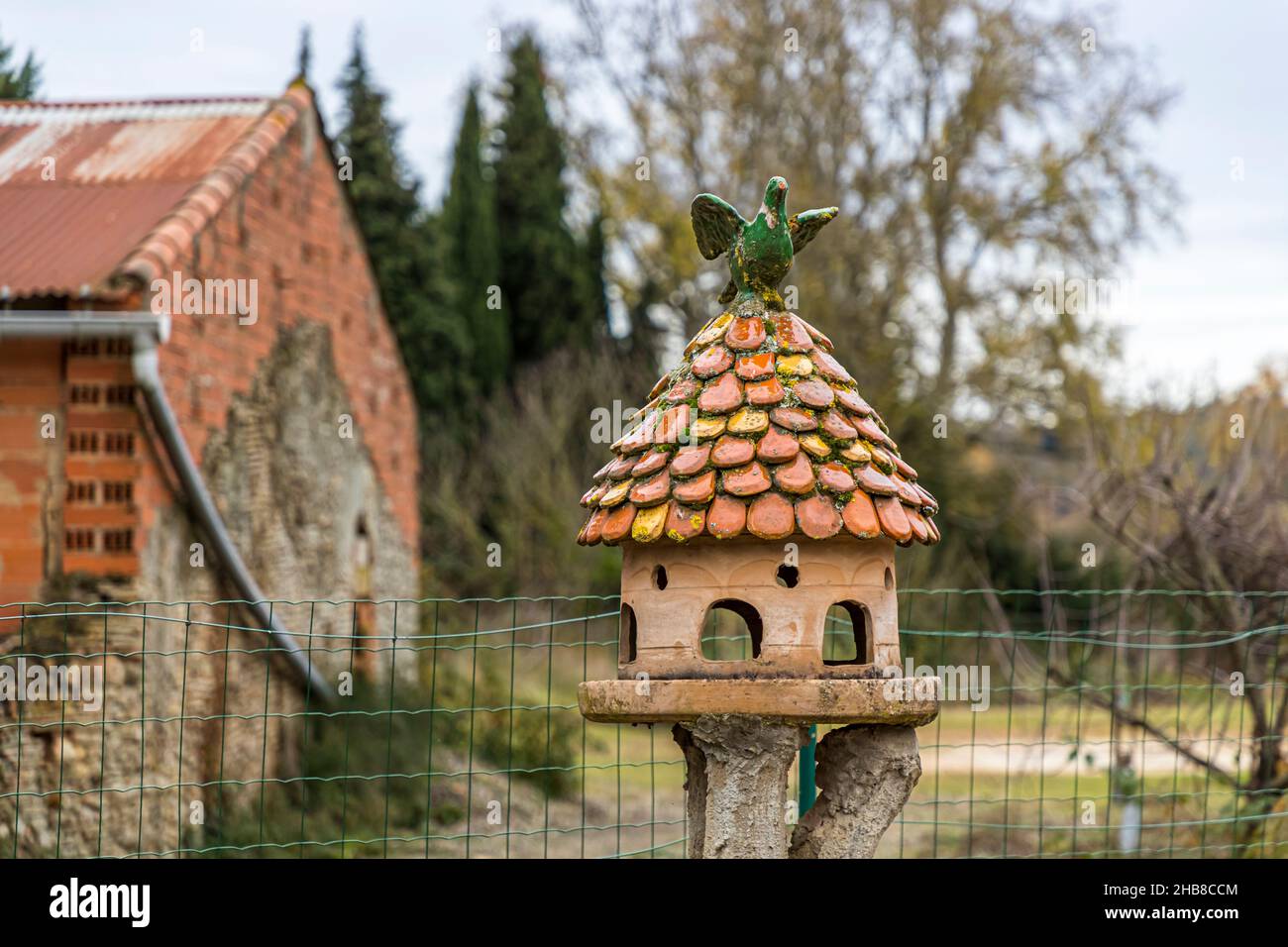 Ein aufwendig verglaster Keramik-Taubenturm steht auf einem Zaunpfahl vor Poterie Not Frères in Mas-Saintes-Puelles, eine grün verglaste Vogelfigur krönt sein schindelgedecktes Dach. Die Werkstatt am Canal du Midi, Département Aude, Frankreich, wurde Anfang des 19. Jahrhunderts von der Familie Perrutel gegründet und wird seit 1947 von der Familie Not betrieben, bekannt für ihre handgefertigte Cassole, das traditionelle Cassoulet-Gericht. Stockfoto