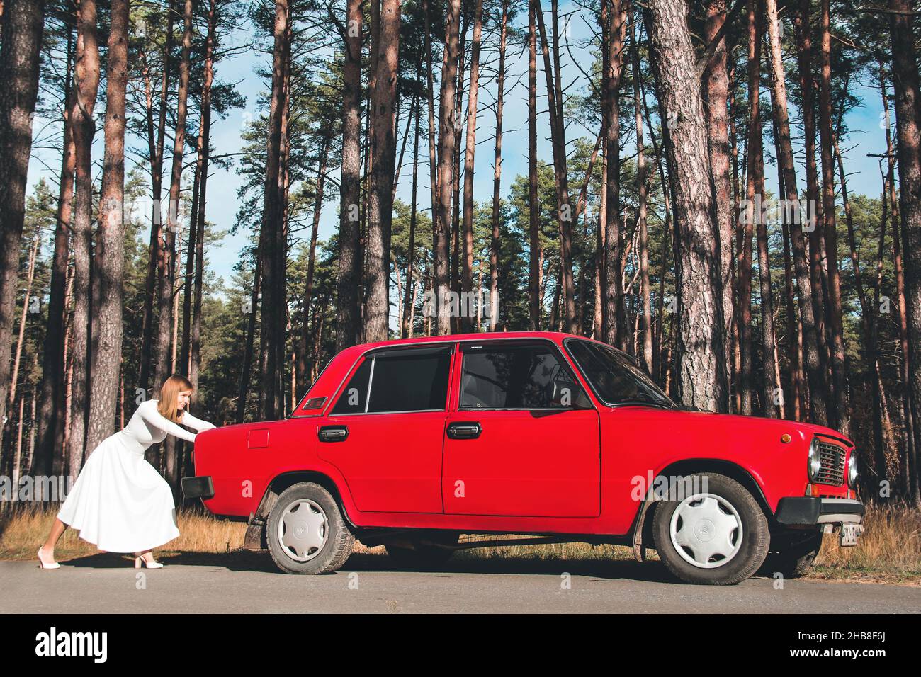 Chernigov, Ukraine - 10. November 2020: Ein Mädchen in einem Hochzeitskleid schiebt ein Auto. Vaz 2101. Rotes Retro-Auto im Wald Stockfoto