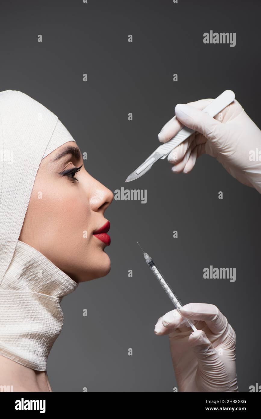 Seitenansicht des Patienten in elastischer Bandage in der Nähe von plastischen Chirurgen mit Operationsmesser und Spritze isoliert auf dunkelgrau Stockfoto