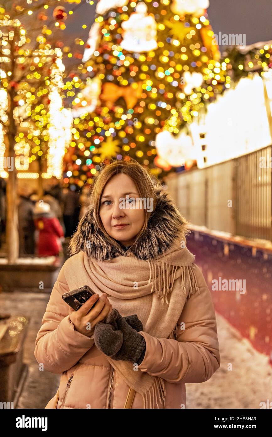 Eine junge Frau auf der Straße, auf dem Platz mit einem Weihnachtsbaum und Weihnachtsdekorationen. Neujahr und Weihnachten feiern Stockfoto