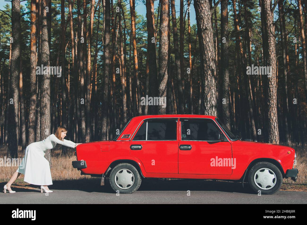 Chernigov, Ukraine - 10. November 2020: Ein Mädchen in einem Hochzeitskleid schiebt ein Auto. Vaz 2101. Rotes Retro-Auto im Wald Stockfoto