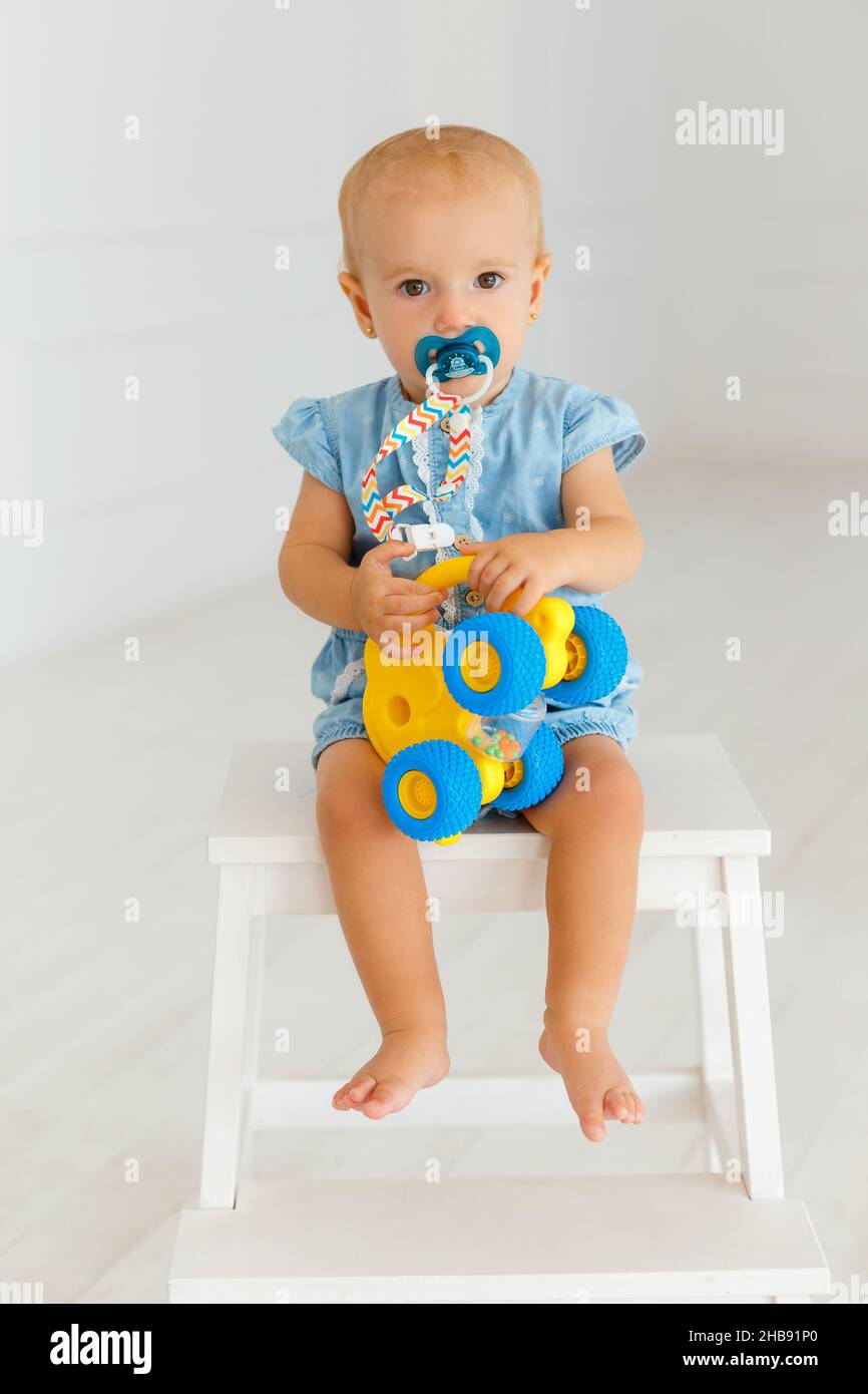 Сute Baby Mädchen in blauen Jeans mit gelben Elefanten. Kleines Kleinkind sitzt auf dem weißen Holzstuhl mit Schnuller im Mund. Porträt eines Jahres ch Stockfoto