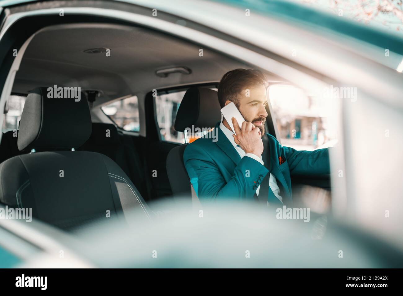 Junger kaukasischer erfolgreicher bärtiger Geschäftsmann in türkisfarbenem Anzug sitzt im Auto und benutzt Smartphone. Versuchen Sie etwas härter, um ein wenig besser zu sein. Stockfoto