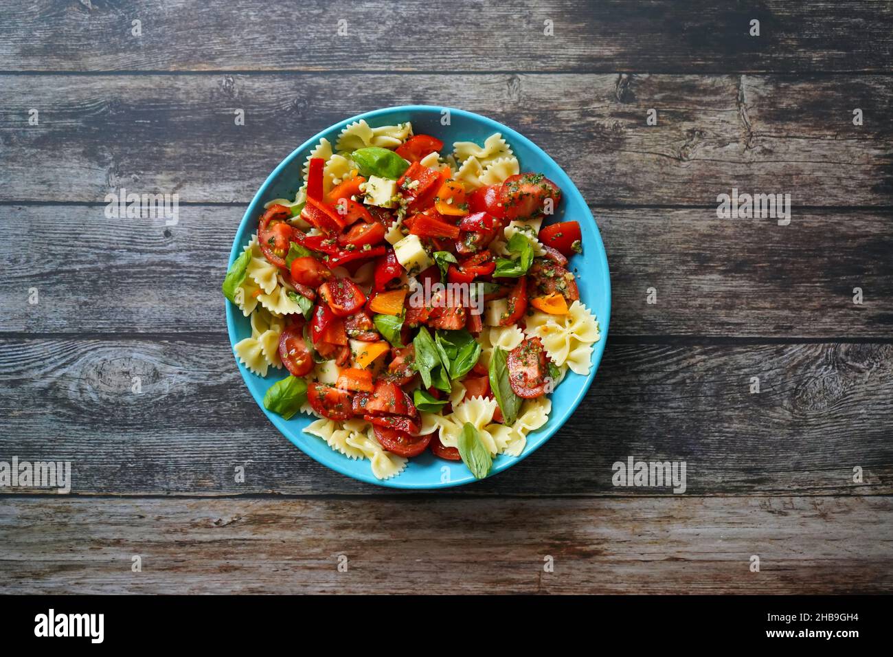 Leichter und saftiger Sommersalat mit Nudeln, Tomaten, Käsewürfeln, Paprika und Basilikum in einer blauen Schüssel. Dunkler Holzhintergrund. Stockfoto