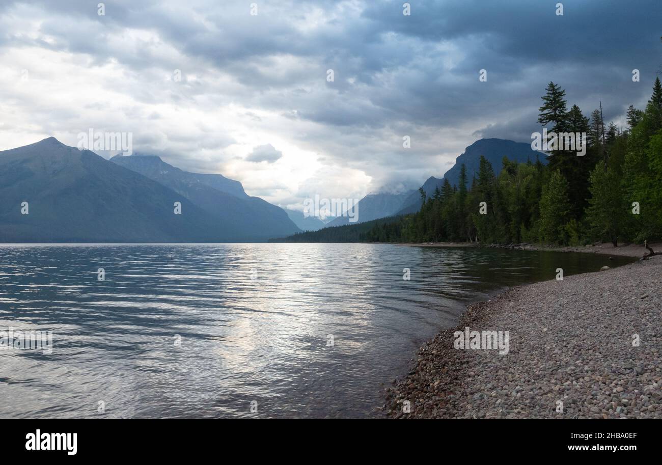 Kiesstrand am Lake McDonald mit immergrünen Wäldern und schroffen Bergen im Hintergrund und Sturmwolken über dem See. Fotografiert in Glacier Nati Stockfoto