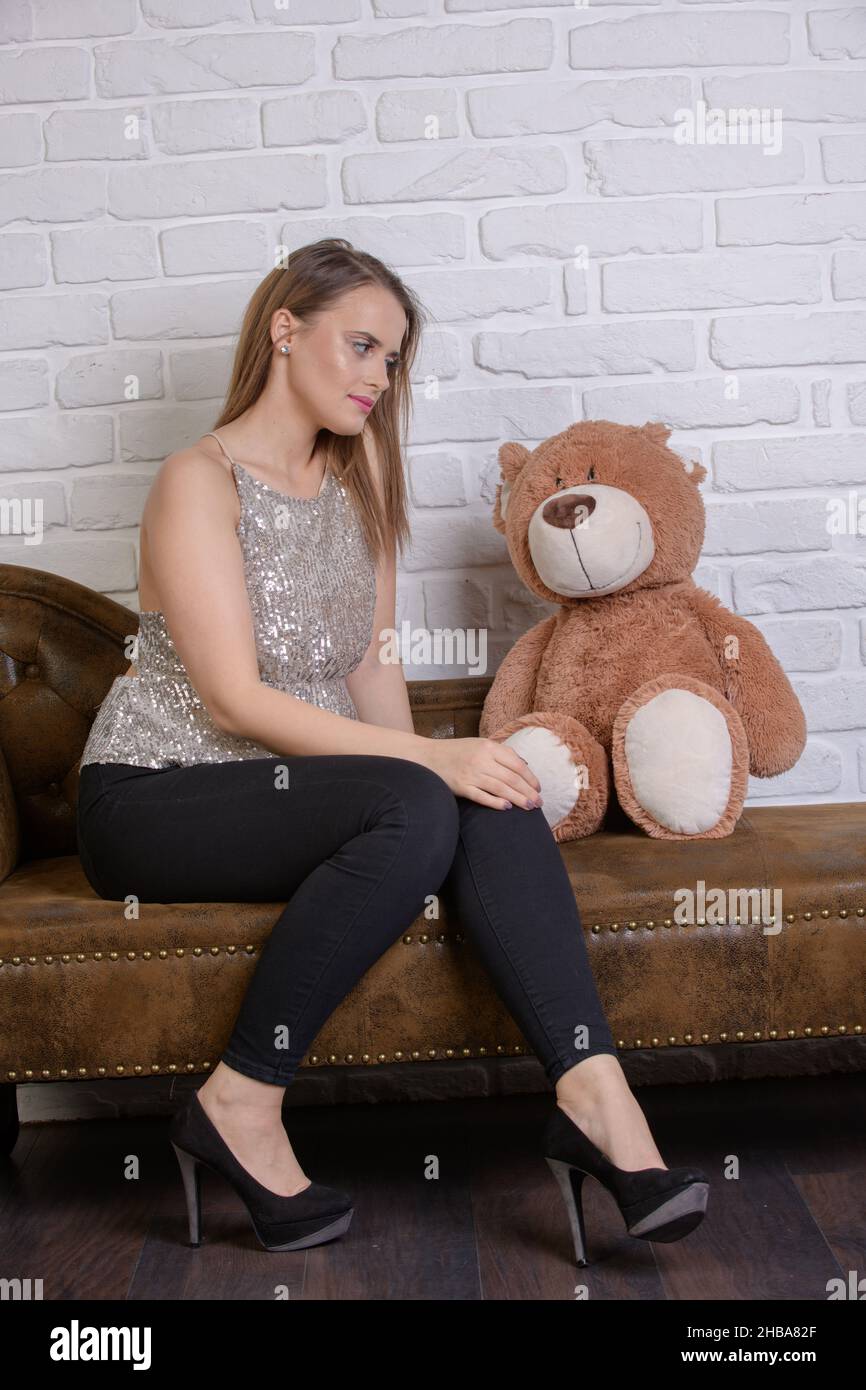 Eine junge hübsche Frau mit langen, geraden Brünetten Haaren und einem großen braunen Teddybär sitzt nebeneinander auf einer Ledercouch. Stockfoto