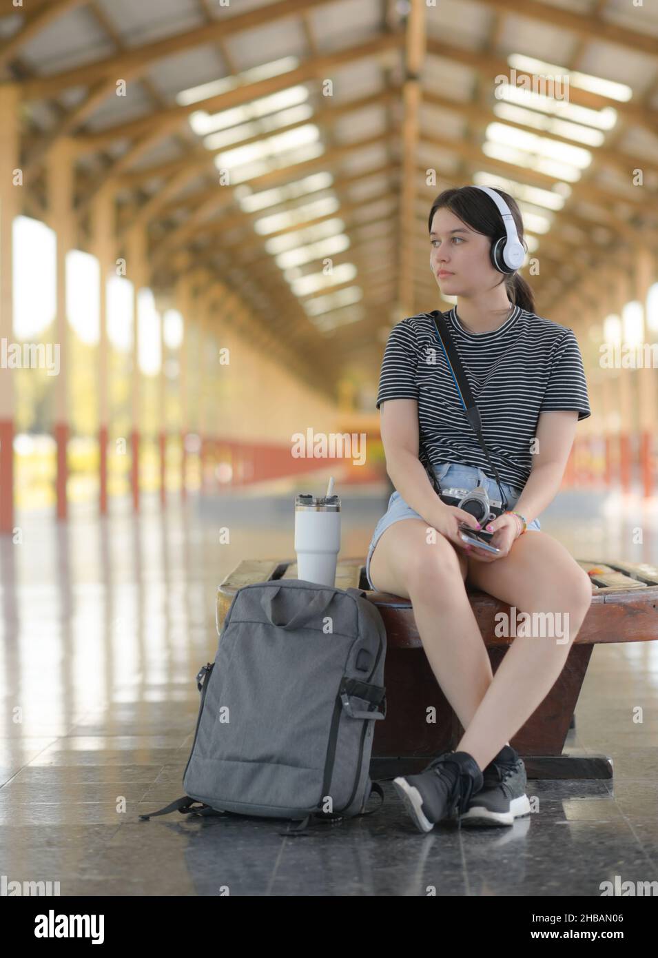 Eine junge Touristenin, die auf einem Bahnsteig Musik hört, eine Kamera trägt und weiße Kopfhörer trägt, um Musik von einer Smartphone-App zu hören Stockfoto