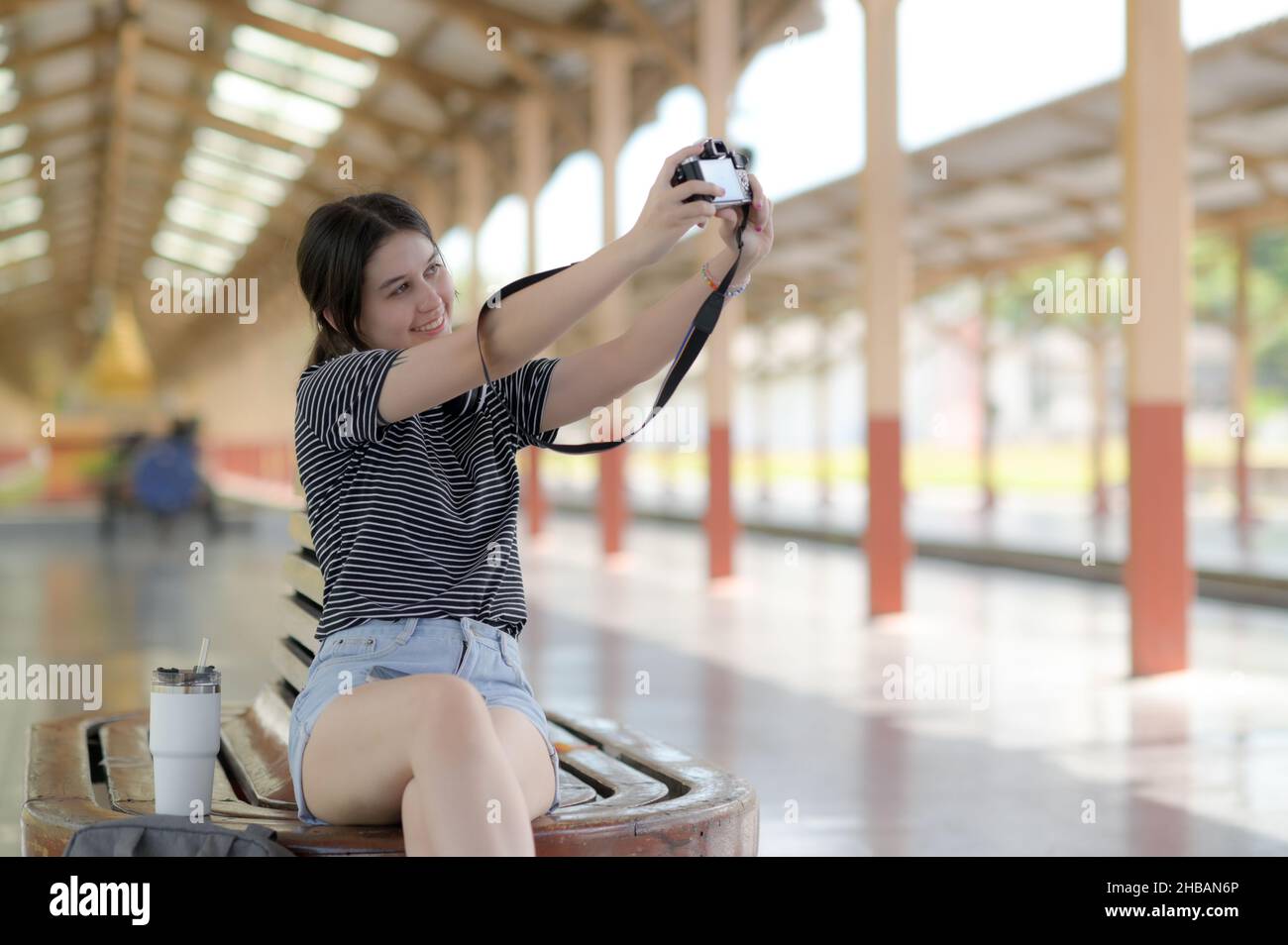 Während sie auf eine Reise wartet, benutzt eine junge Touristenin eine Selfie-Kamera, ausländische Jugendliche Mädchen machen Selfies, ein weißer Reisetasse wird auf den Chai gelegt Stockfoto