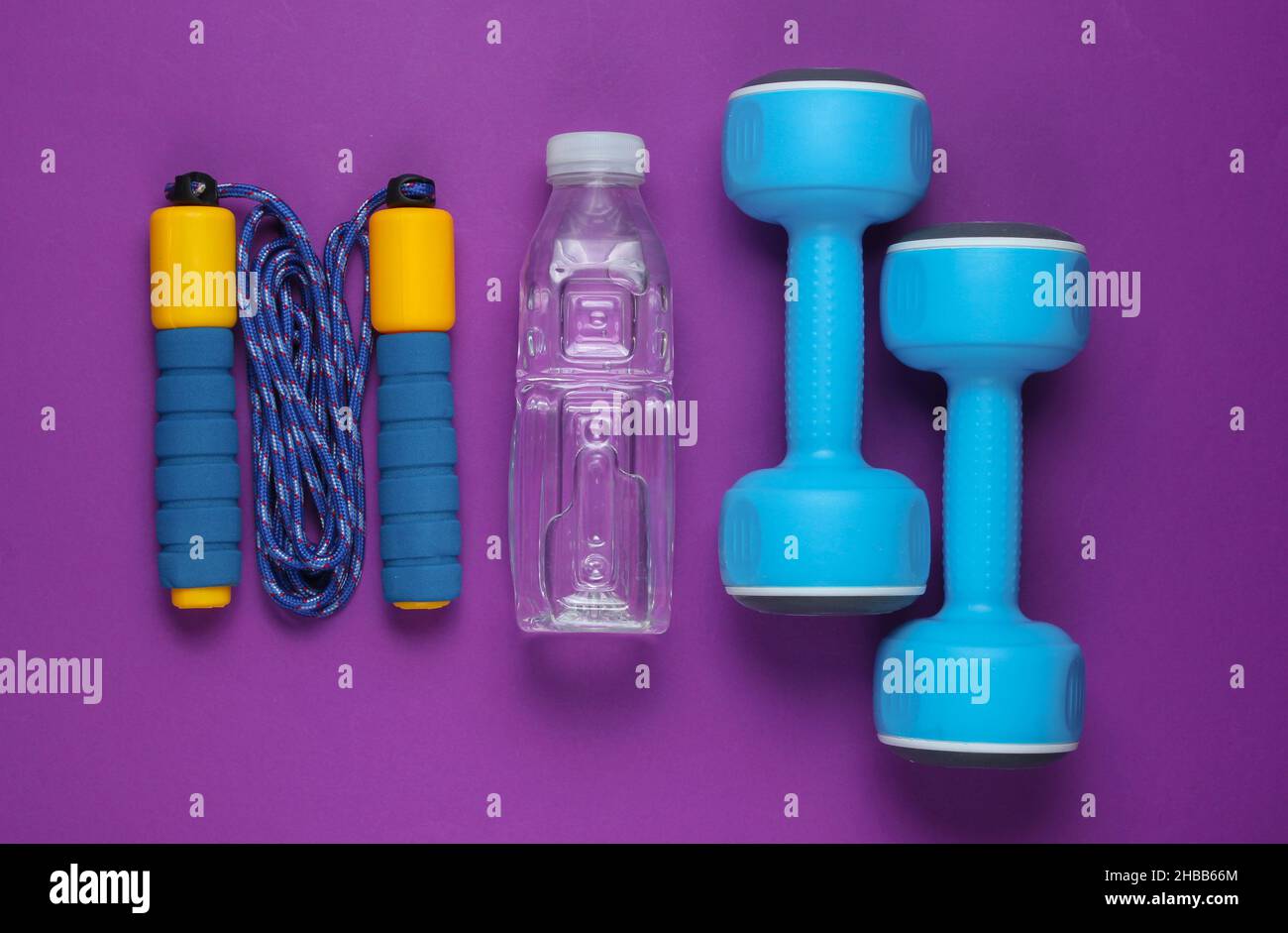 Sportkonzept im flachen Lay-Stil. Kurzhanteln, Springseil, Flasche Wasser. Sportgeräte auf violettem Hintergrund. Draufsicht Stockfoto