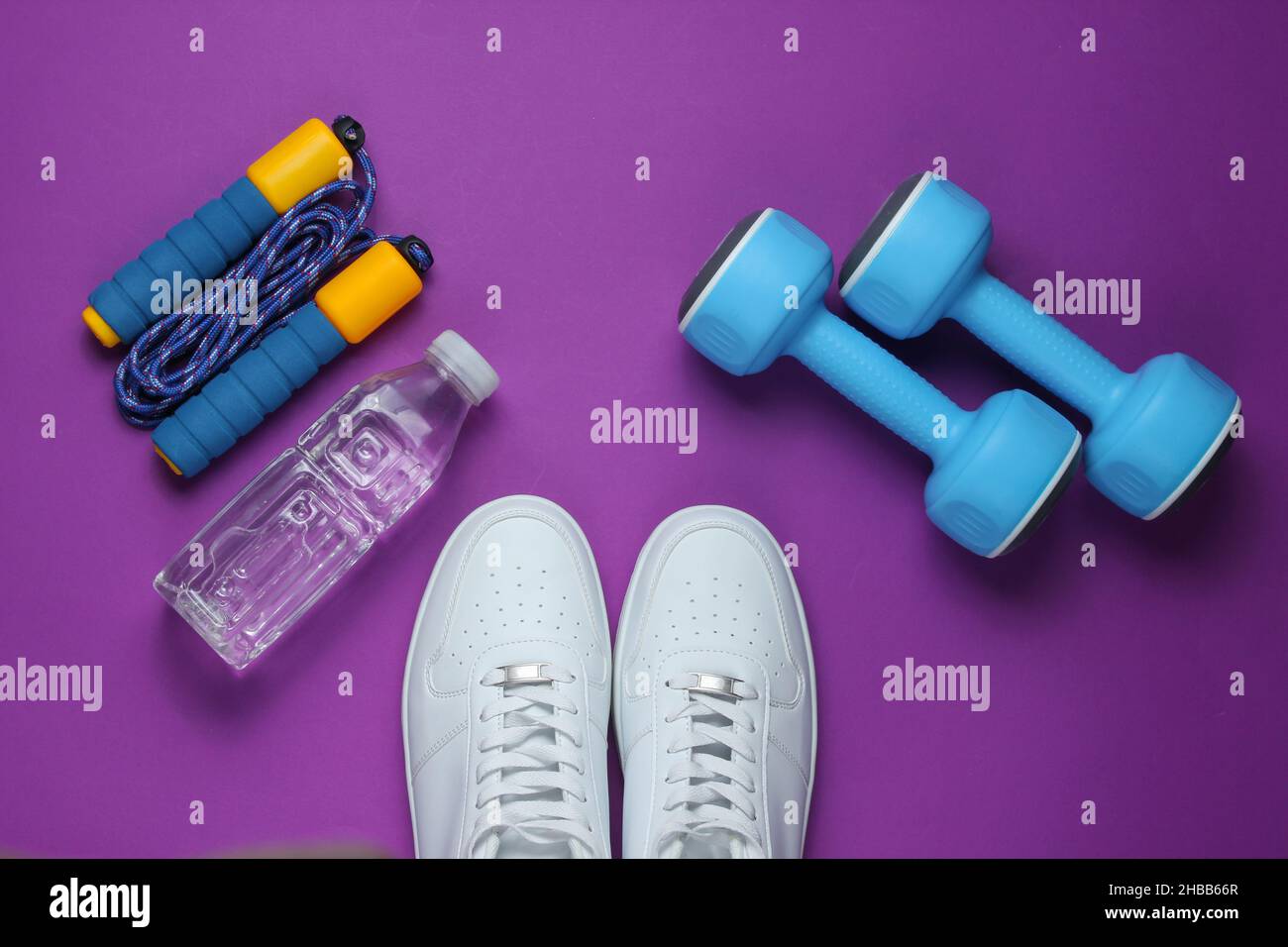 Sportkonzept im flachen Lay-Stil. Kurzhanteln, Turnschuhe, Springseil, Flasche Wasser. Sportgeräte auf violettem Hintergrund. Draufsicht Stockfoto