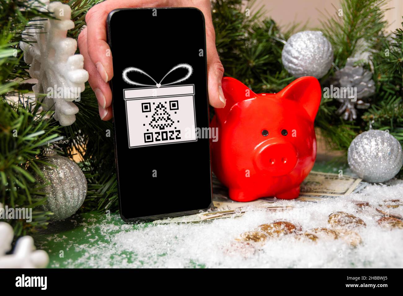 Eine Hand, die ein Smartphone mit einem Geschenksymbol und einem QR-Code in Form einer Tanne hält, ein rotes Sparschwein auf mit Schnee und Christma bedeckten Goldmünzen Stockfoto