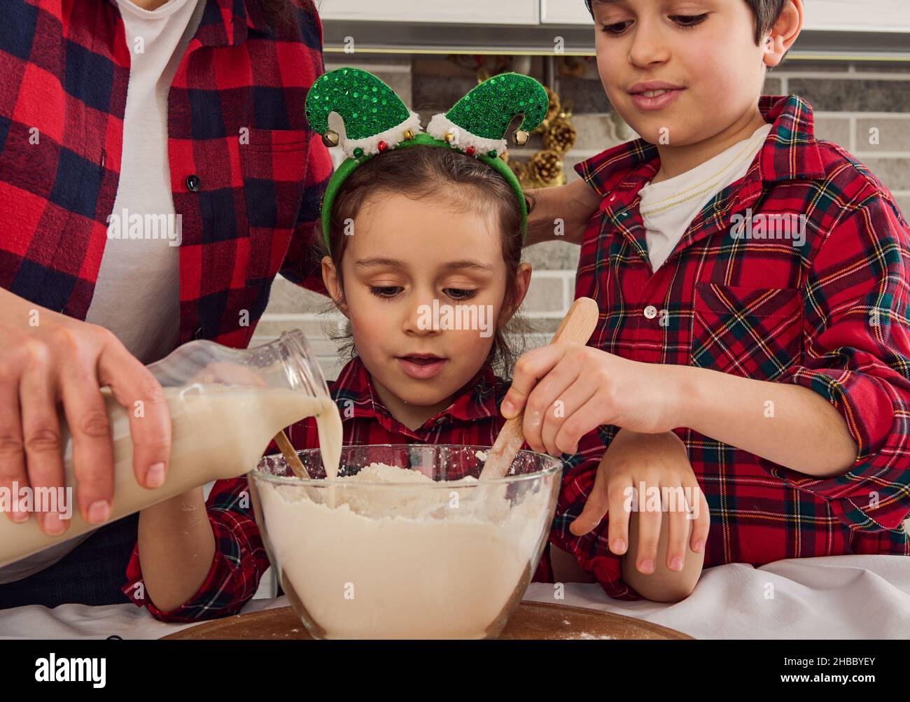 Das hübsche kleine Mädchen sitzt zwischen ihrer liebenden Mutter, die etwas Milch in eine Schüssel mit Mehl gießt, und ihrem fröhlichen älteren Bruder, der trocken mischt und knetet Stockfoto