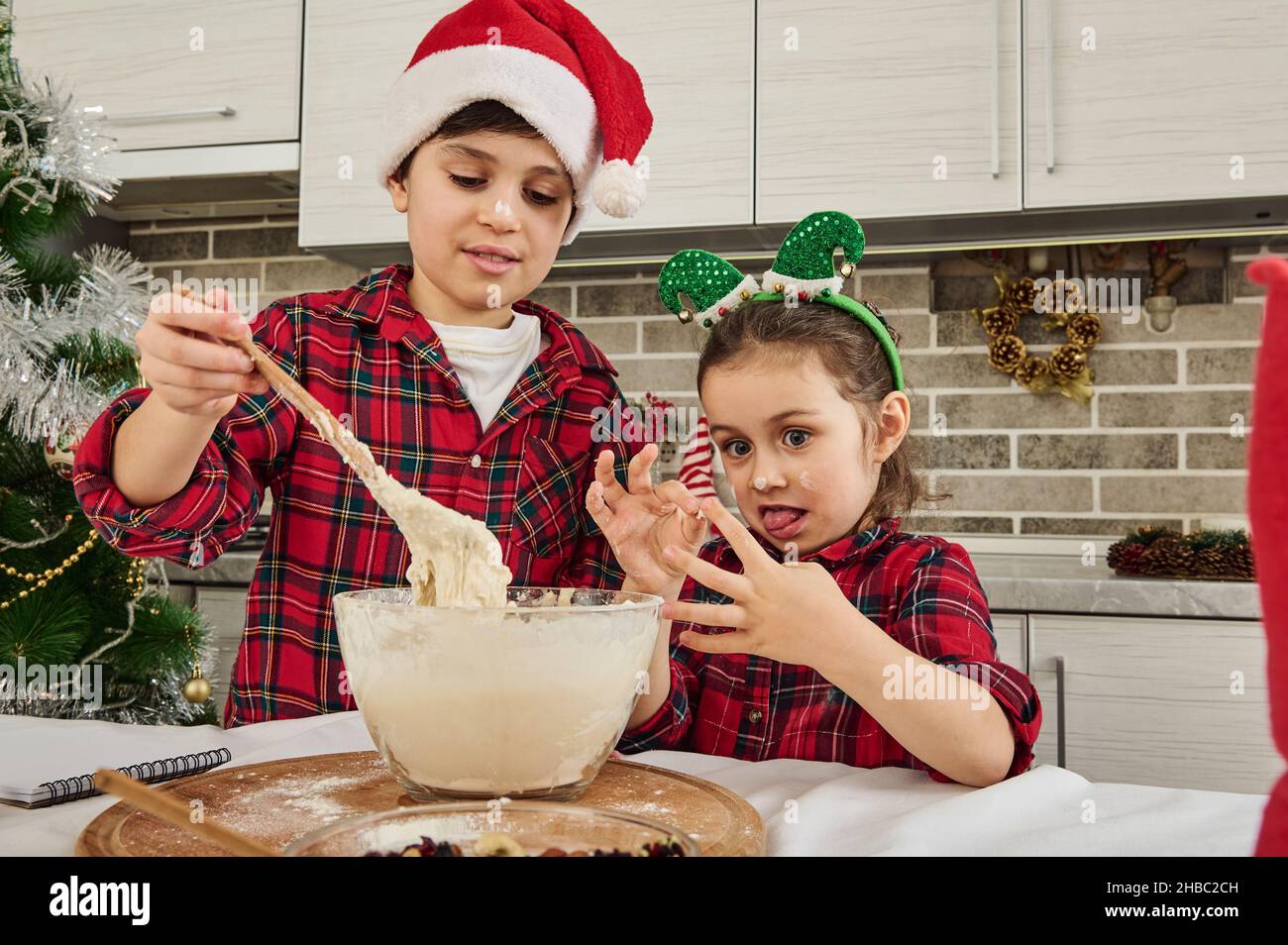 Entzückende europäische Kinder, die zusammen Spaß haben und Teig für Weihnachtskekse, Lebkuchen, Muffins in der heimischen Küche zubereiten. Bruder in Santa hat Stockfoto