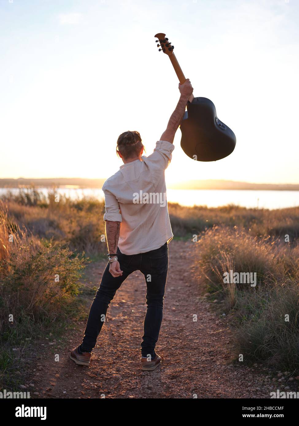 Gitarrist posiert mit seiner Gitarre hoch in der Luft vor einem Sonnenuntergang an einem See Stockfoto