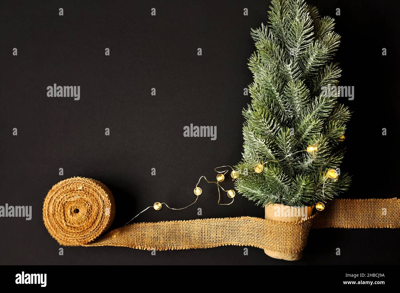 Vorbereitung von Dekorationen für Winterfeste. Künstlicher Weihnachtsbaum, natürlicher Jutefaserstreifen, dunkler Hintergrund, gemütliche Beleuchtung Stockfoto