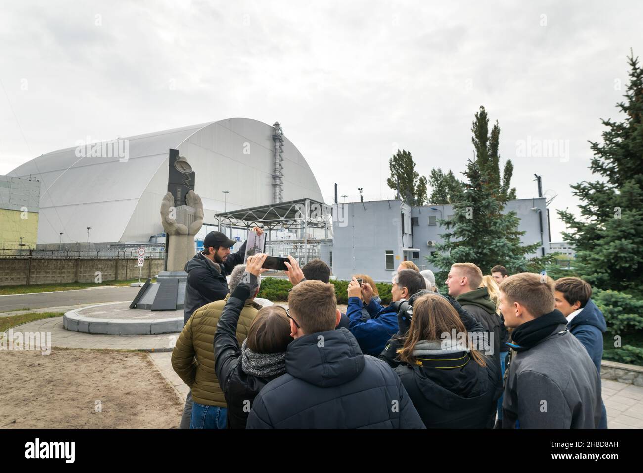 Männlicher Führer vor der Gedenkstätte von Tschernobyl zeigt ein Foto des Kernkraftwerks nach einem Unfall.das Kernkraftwerk Tschernobyl steht im Hintergrund Stockfoto