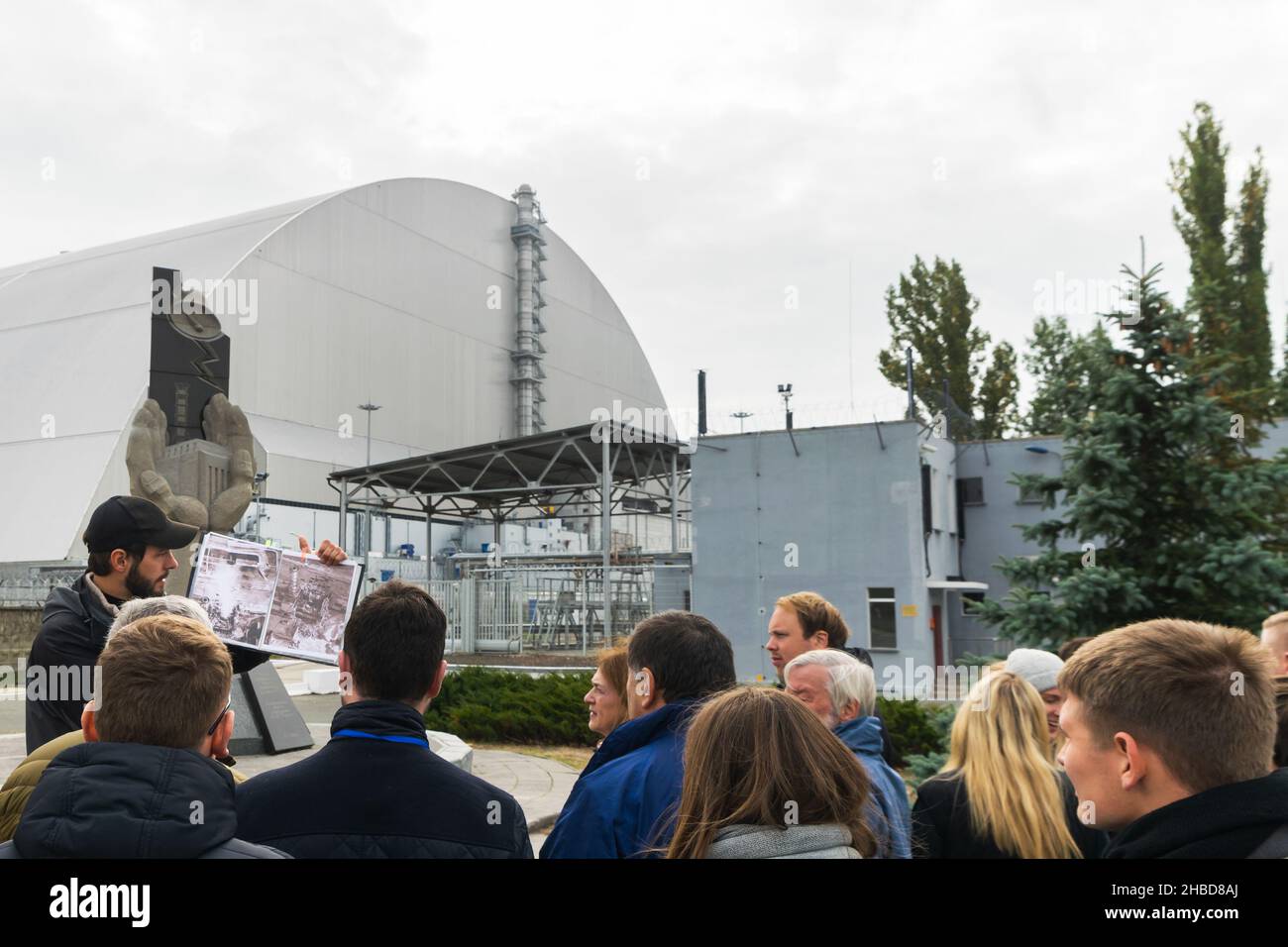 Männlicher Führer vor dem Tschernobyl-Denkmal zeigt ein Foto des Atomreaktorblocks nach der Explosion. Das Kernkraftwerk Tschernobyl steht in t Stockfoto