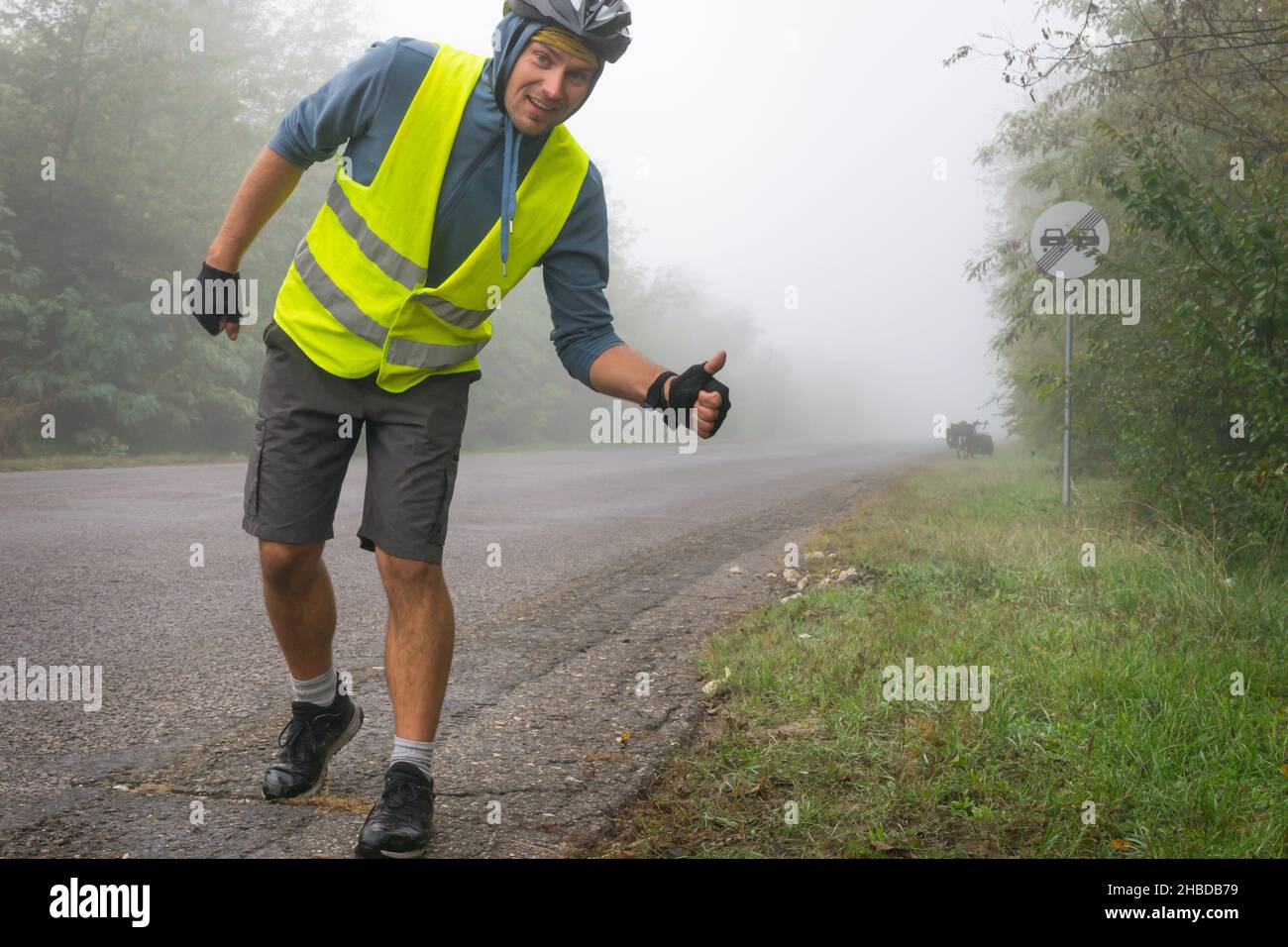 Ein glücklicher Radfahrer mit einer reflektierenden Weste zeigt bei nebligen Bedingungen die Daumen nach oben auf der Straße mit einem Touring-Fahrrad in der Nähe. Konzept für sicheres Radfahren unter b Stockfoto