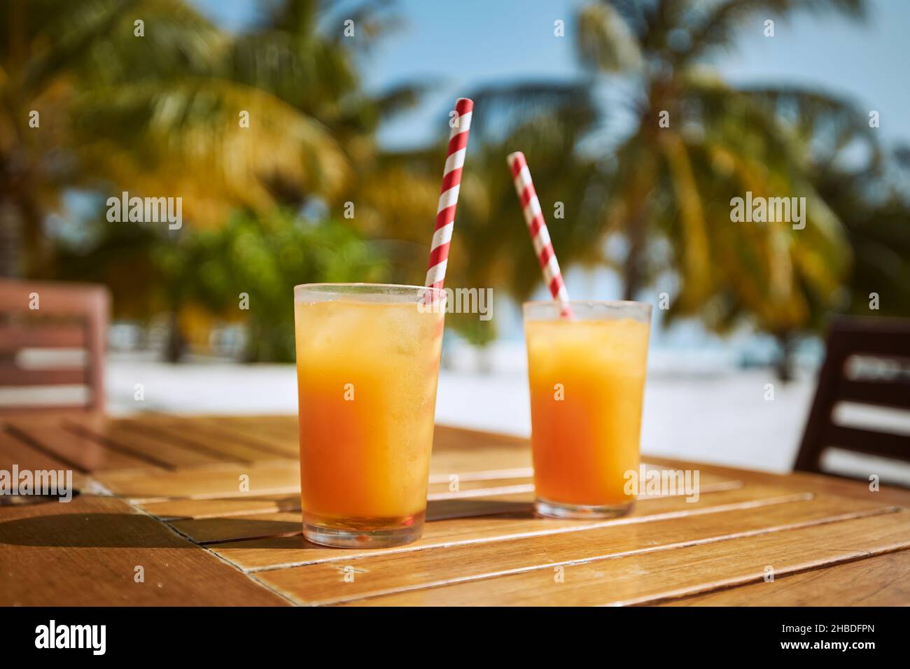 Zwei erfrischende kalte Getränke am weißen Sandstrand. Gläser Cocktails mit Trinkhalmen aus Papier auf einem Holztisch gegen Palmen. Stockfoto