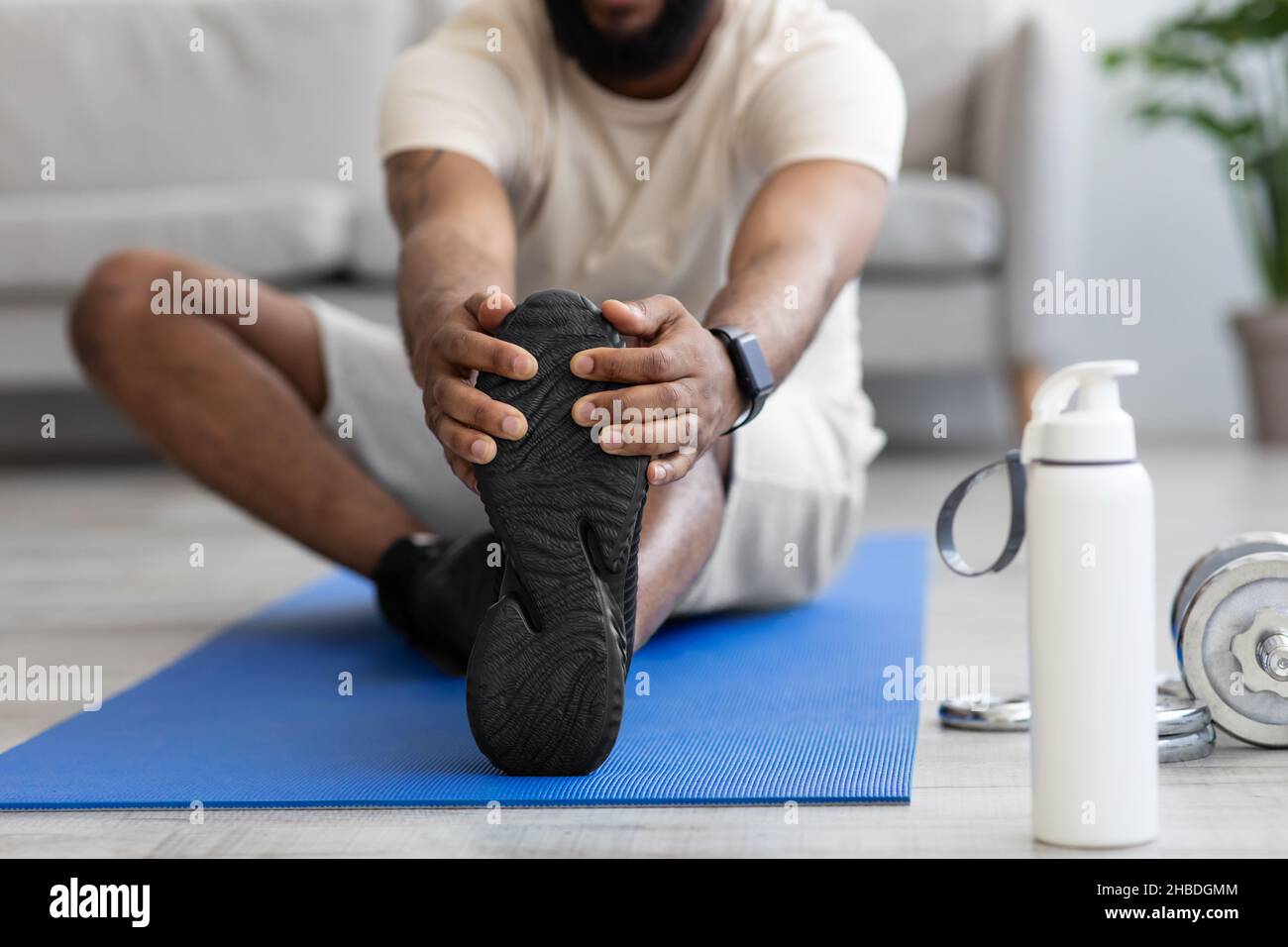 Junger schwarzer Mann macht auf der Matte mit Hanteln und einer Flasche Wasser Übungen für Beine oder Stretching Stockfoto