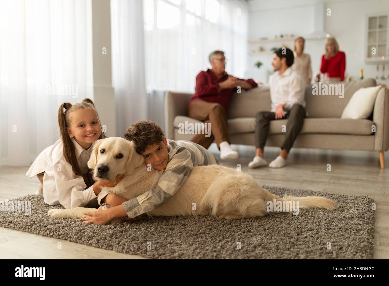 Glückliche Kinder umarmen ihren goldenen Retriever, liegen auf dem Boden zusammen, ihre Verwandten kommunizieren auf der Couch zu Hause Stockfoto