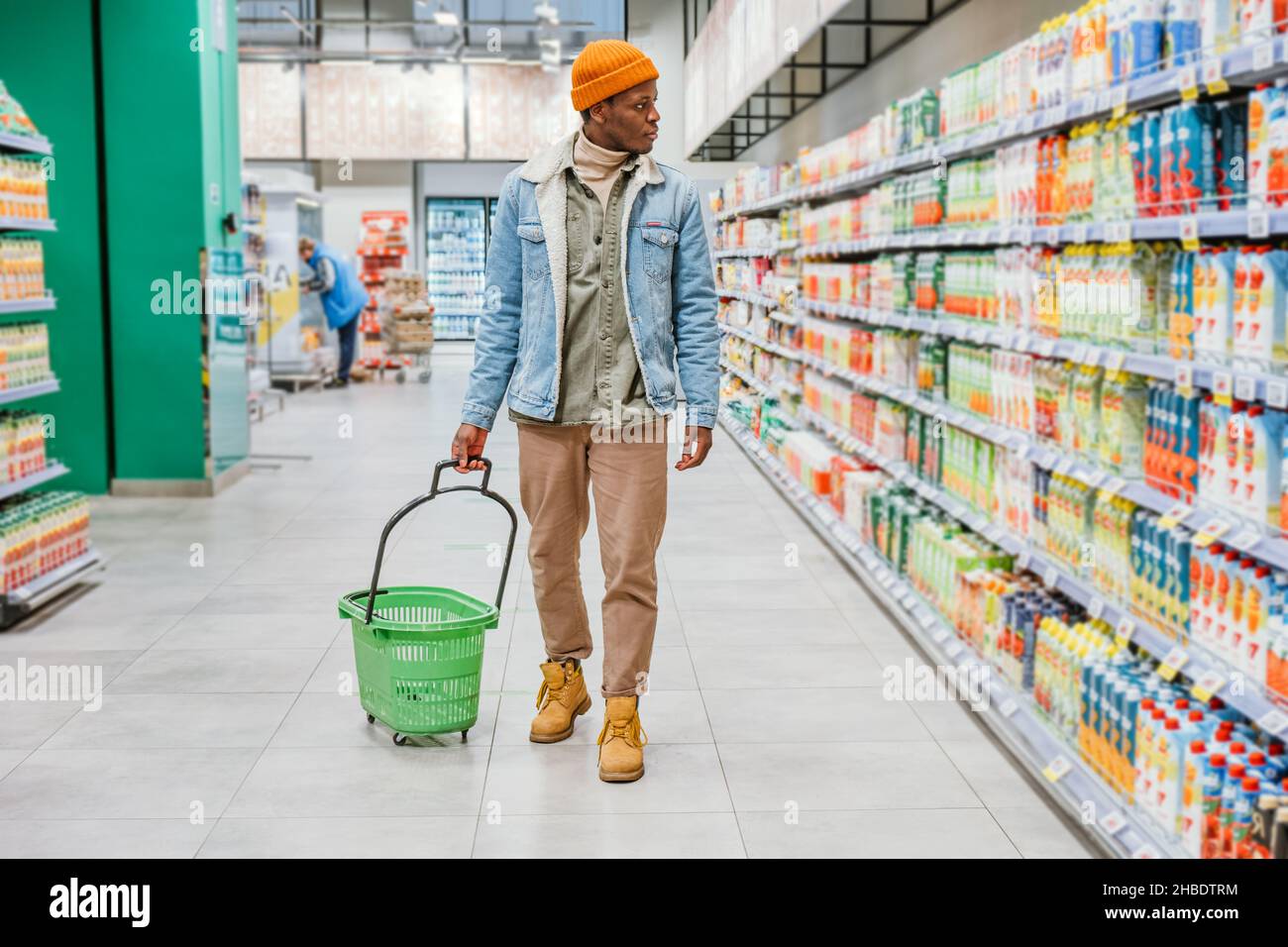 Afroamerikanischer Mann in einem stilvollen orangefarbenen Hut geht mit einem leeren grünen Einkaufswagen durch einen Supermarkt und wählt leckeres und gesundes Essen für ein Familienessen. Stockfoto