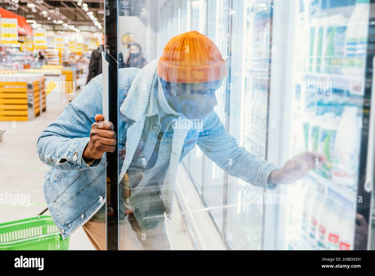 Afrikanischer Hipster-Mann öffnet die Glastür eines Kühlschranks in einem Supermarkt und wählt frische abgefüllte Milch oder Kefir und Joghurt Stockfoto