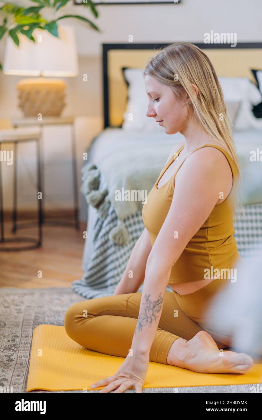 Die junge blonde langhaarige Frau in gelber Sportkleidung meditiert Yoga und sitzt in Stretching-Pose mit geschlossenen Augen auf der Matte im Schlafzimmer Stockfoto