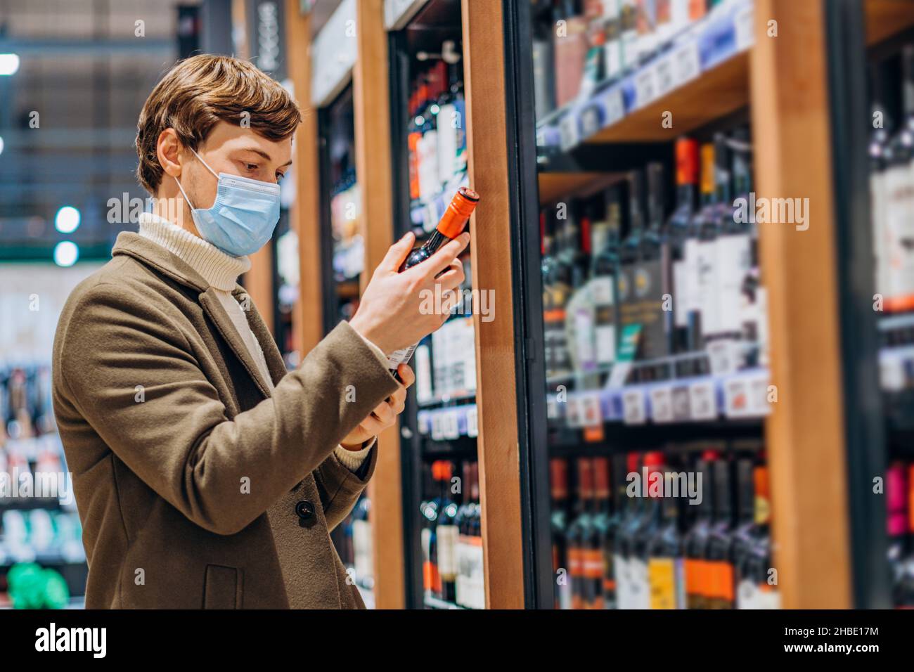 Junger Mann mit welligen Brunet Haar trägt medizinische Maske Pullover und warmen Mantel wählt eine Flasche Wein für Feiertagsfeier Partei Stockfoto
