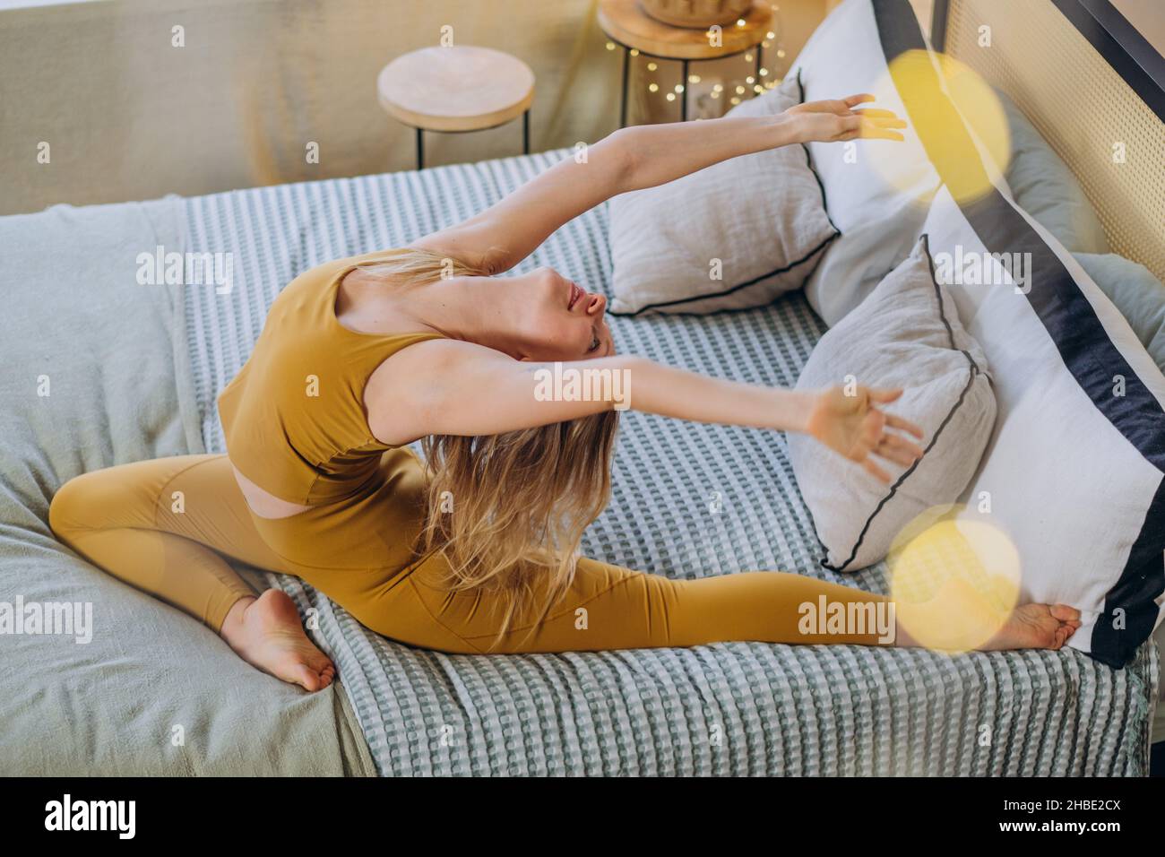 Die junge blonde Frau trägt ein Sporttop und Leggings, die Yoga-Posen machen, und dehnt sich auf einer weichen Decke, die das Bett mit Girlande-Beleuchtung bedeckt Stockfoto