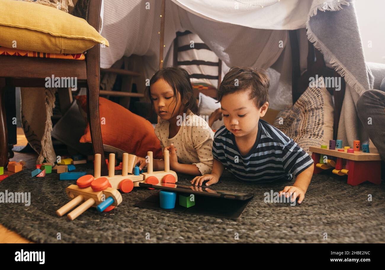 Zwei junge Kleinkinder spielen zuhause zusammen. Zwei entzückende junge Geschwister, die mit Spielzeug spielen und Kinder auf einem digitalen Tablet ansehen. Sibl Stockfoto