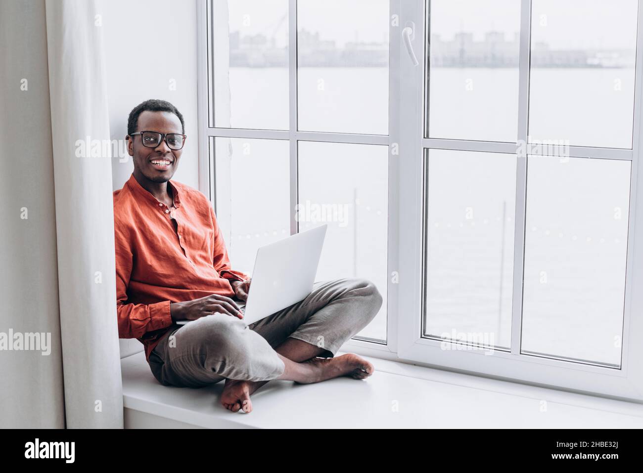 Ein junger afrikanischer Geschäftsmann mit Brille sitzt auf einem Laptop auf dem Fensterbrett eines Hauses und schaut lächelnd auf die Kamera Stockfoto