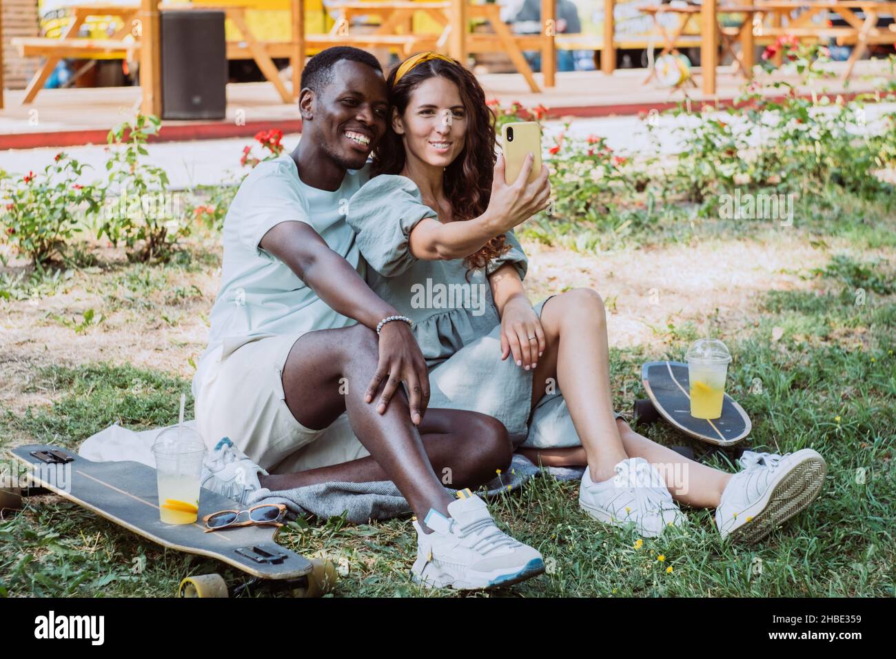 Der junge schwarze Mann und die weiße Brünette machen Selfie auf dem Smartphone, das auf dem Bettüberwurf mit Skateboards sitzt und im Park auf Gras trinkt Stockfoto