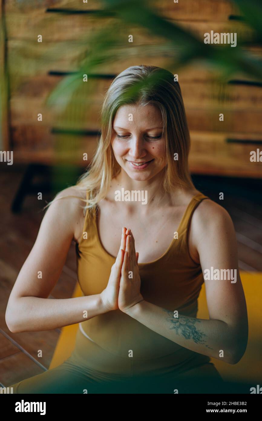 Eine athletische Frau mit blonden, lockeren Haaren und Tätowierungen in der Oberseite und Leggings meditiert mit einem breiten Lächeln in Lotushaltung auf der Yoga-Matte aus der Nähe Stockfoto