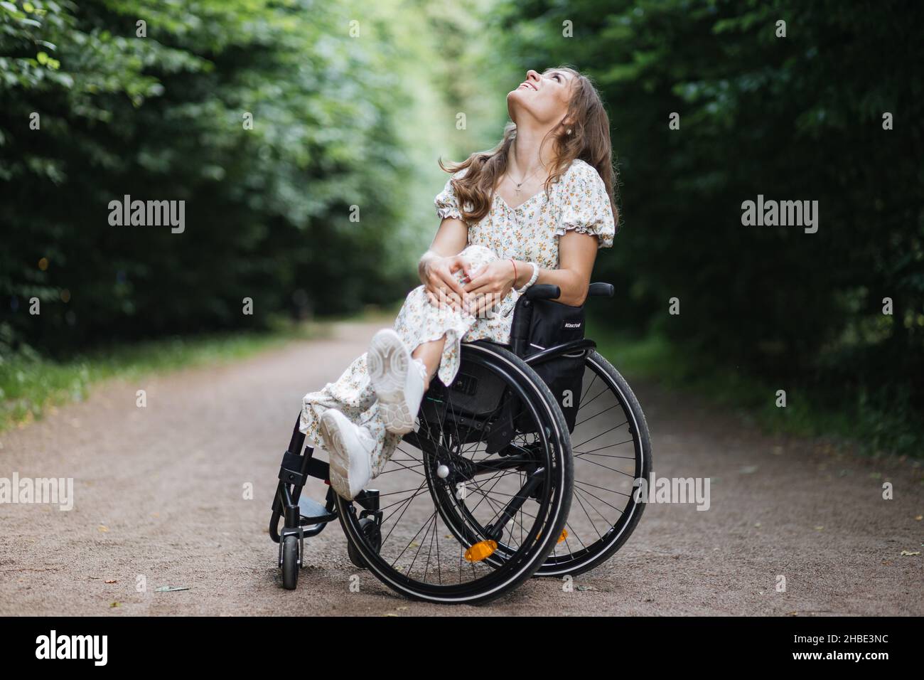 Attraktive kaukasische Frau im Sommerkleid sitzt im Rollstuhl zwischen grünen Park und schaut nach oben. Weibliche Person mit körperlicher Behinderung genießen jeden Moment im Freien. Stockfoto