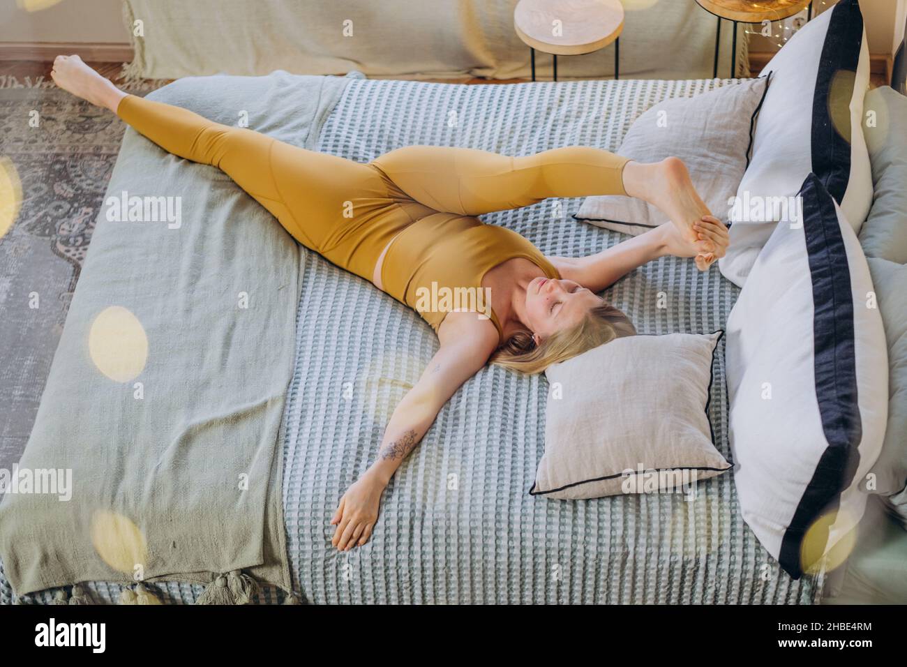 Athletische Frau mit blonden Haaren in gelber Sportswear streckt sich, spaltet sich und liegt auf einer weichen Decke, die das Bett mit Kissen aus der oberen Ansicht bedeckt Stockfoto