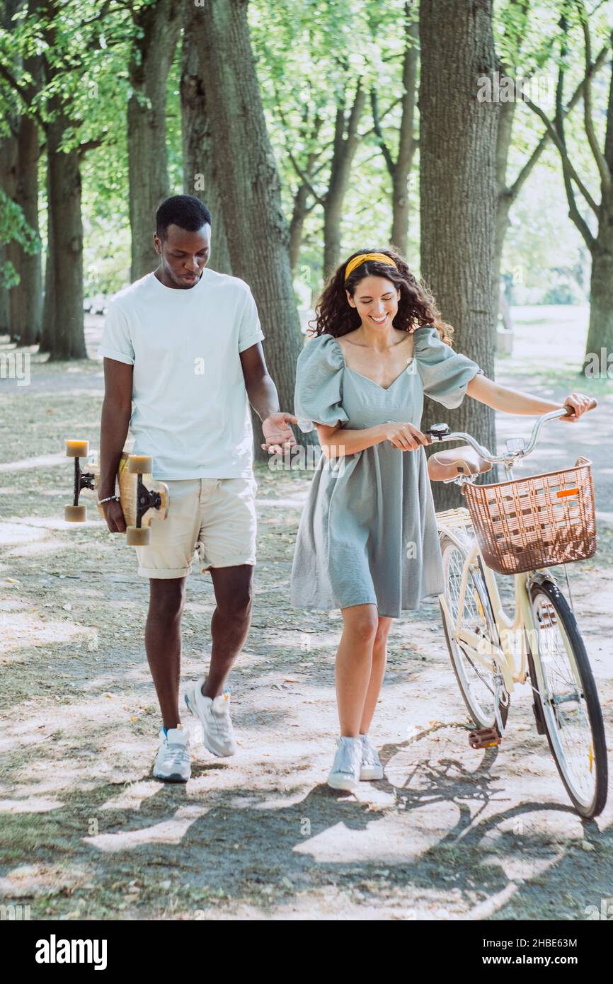 Afroamerikanischer junger Mann mit Skateboard und brünette Dame mit Fahrradtour und Lächeln im Sommerpark am sonnigen Nachmittag Stockfoto
